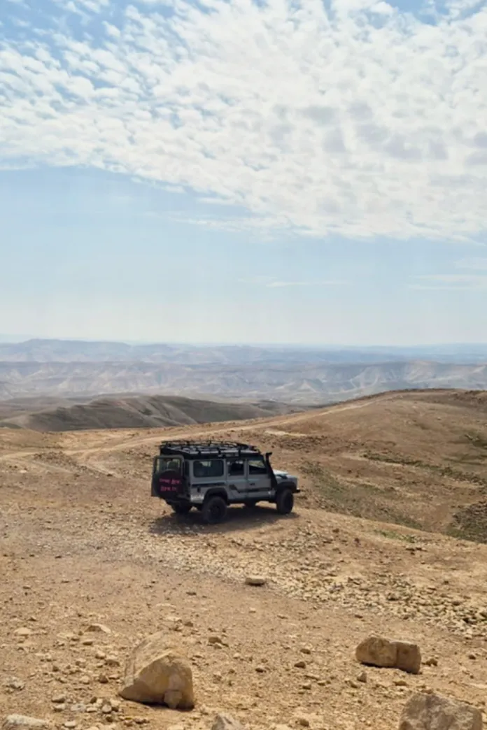 Four-wheel drive vehicle traveling through the Judean Desert near the Dead Sea, surrounded by rolling desert hills beneath a wide, cloud-filled sky.