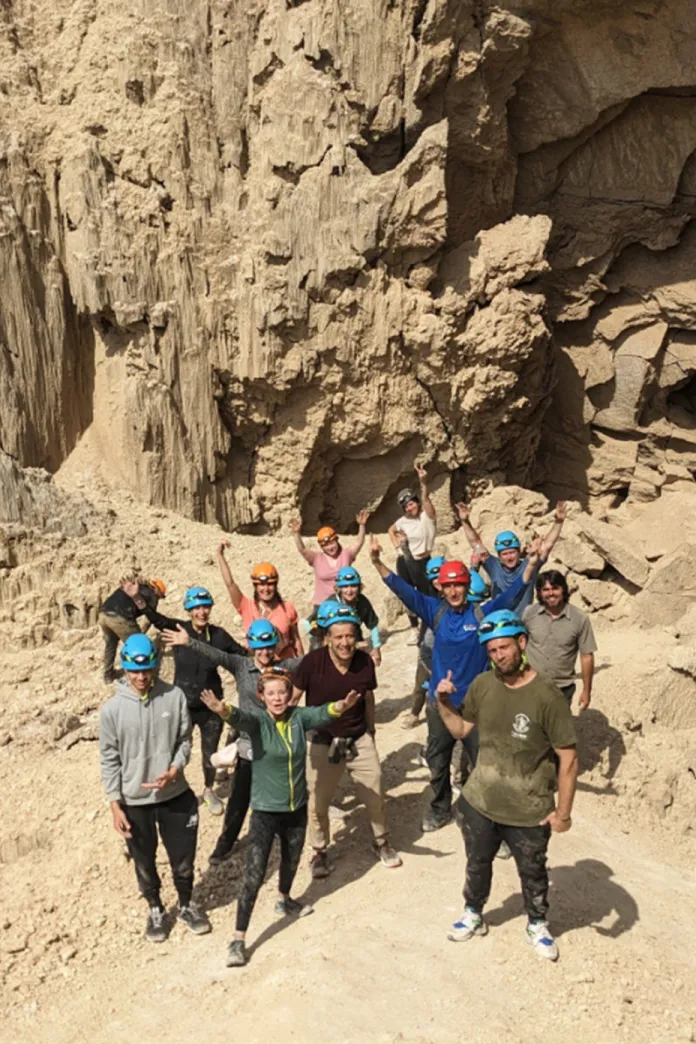 Group of adventurers wearing helmets posing at the entrance to Malcham Cave near the Dead Sea, surrounded by towering desert rock formations.