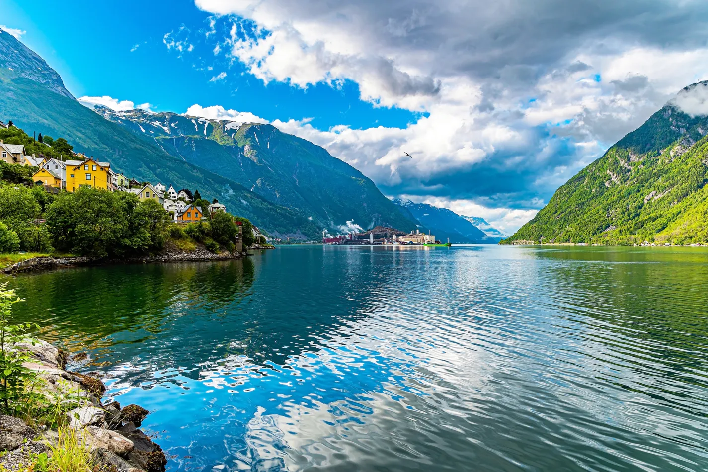 Vibrant blue sky and towering green mountains with colorful houses nestled in the trees along the water's edge