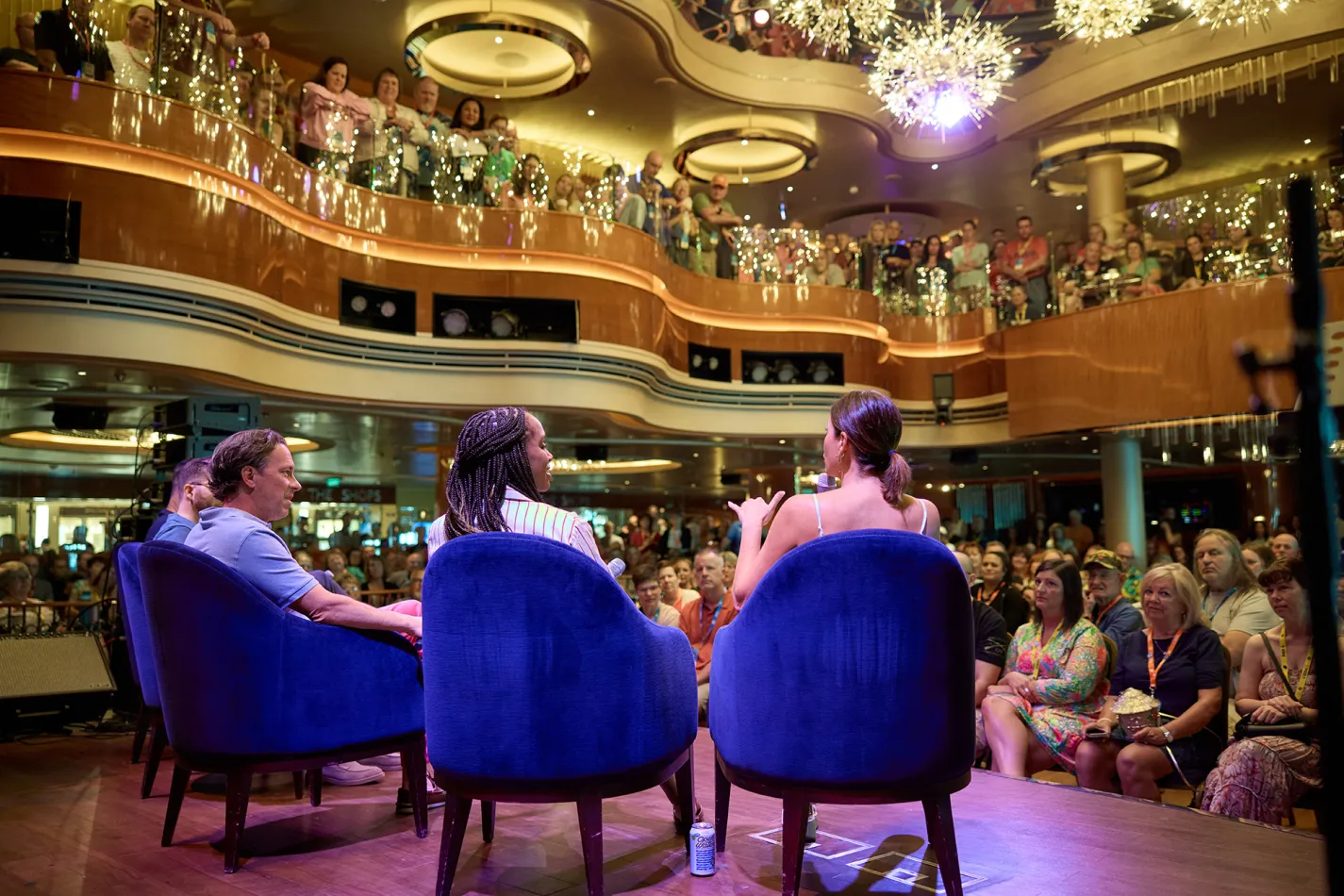 Speakers on a stage, sitting in blue chairs, talking to a room full of people