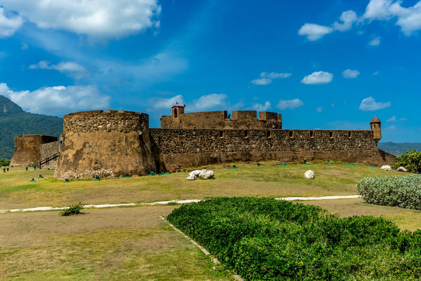 Historic stone fortress in Puerto Plata with thick defensive walls and watchtowers under a bright blue sky with scattered clouds, surrounded by grassy grounds and low shrubs.