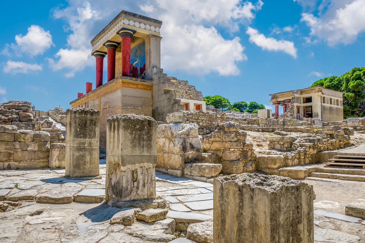 Ruins of the ancient Palace of Knossos in Crete, featuring restored red columns, stone pathways, and archaeological remains under a bright blue sky.