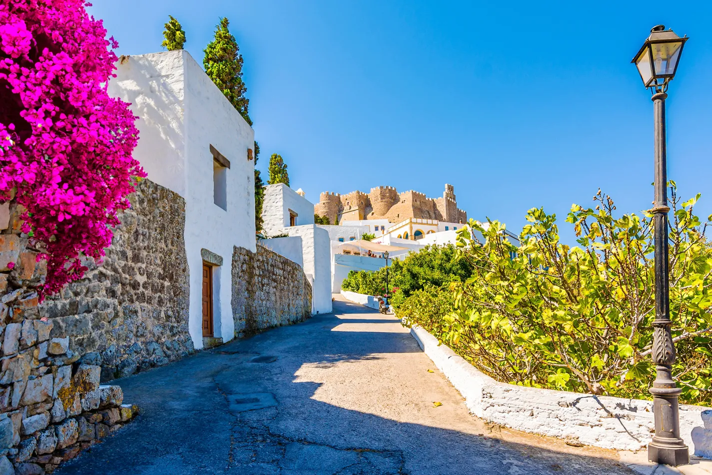Whitewashed buildings and colorful bougainvillea line a quiet street in Patmos, with the historic Monastery of Saint John visible on the hillside above.