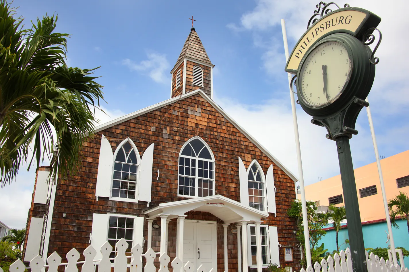 Historic wooden church in Philipsburg, St. Maarten, with a nearby street clock, white picket fence, and palm trees beneath a blue, cloud-filled sky.