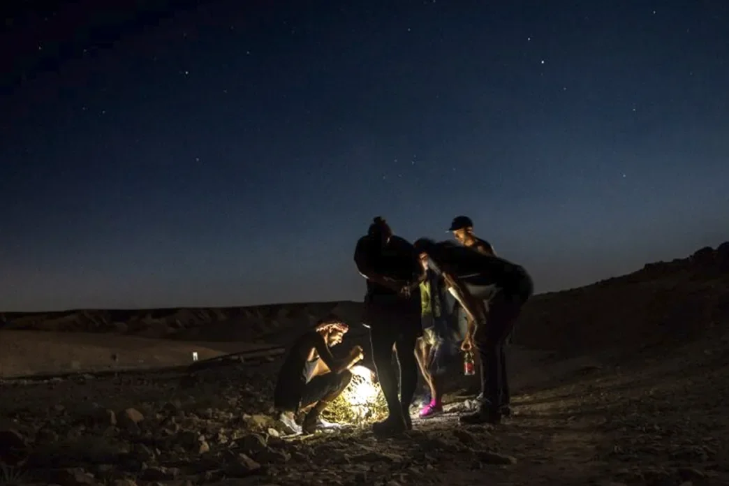 A small group gathered around a light source in the desert under a starry night sky
