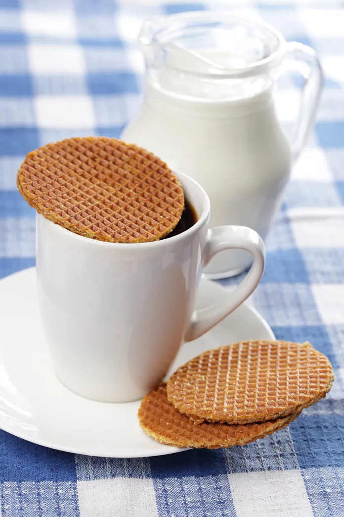 A white coffee cup with waffle cookies on it and a small pitcher of milk next to it
