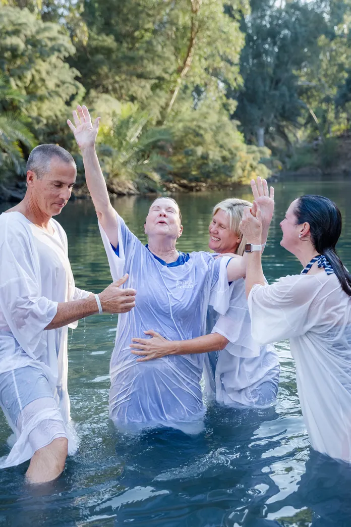 Woman in white robe raises her arms in joy while being baptized in the Jordan river
