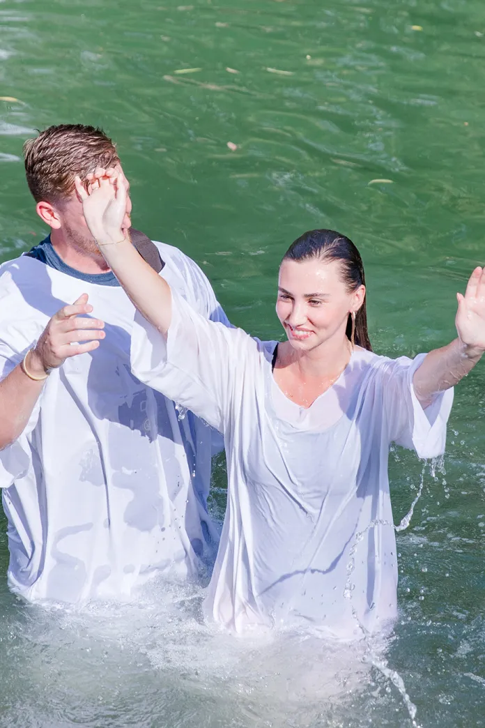 A woman in a white robe smiles and raises her hands while standing in a body of water during a baptism ceremony, with another participant beside her.