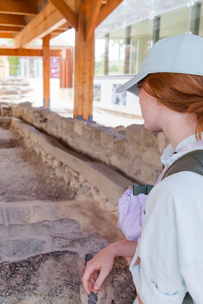 A person wearing a light-colored cap and backpack looks down at uncovered stone ruins beneath a wooden shelter.