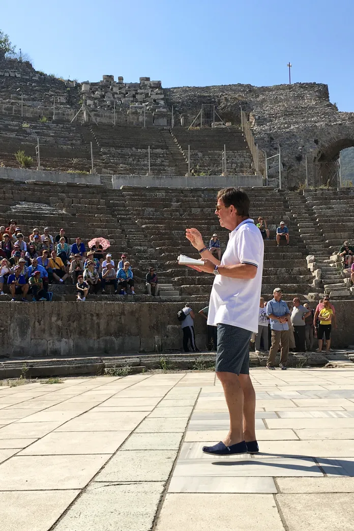 Alistair Begg speaking to a group seated in the ancient theater at Ephesus, surrounded by historic stone ruins and tiered seating.