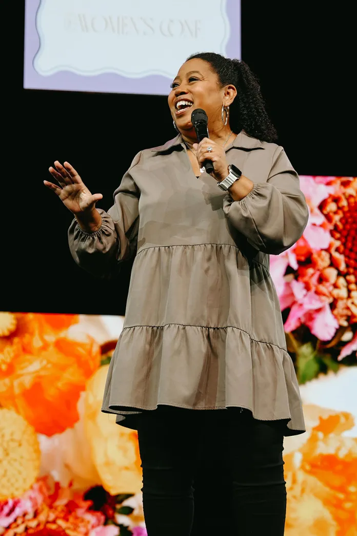 Chrystal Hurst speaking on stage with microphone, smiling; bright orange floral backdrop