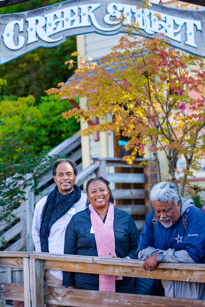 Three people smiling on a wooden balcony under Creek Street sign, autumn trees behind.