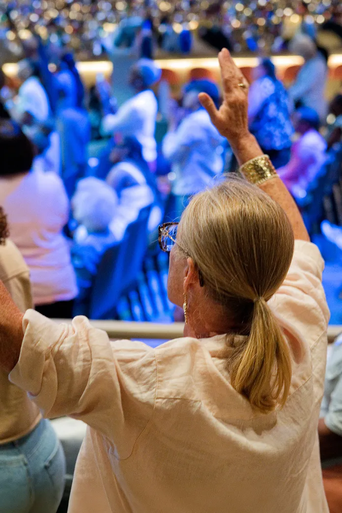 Woman with blonde ponytail raises hands in a blue-lit crowd at an event
