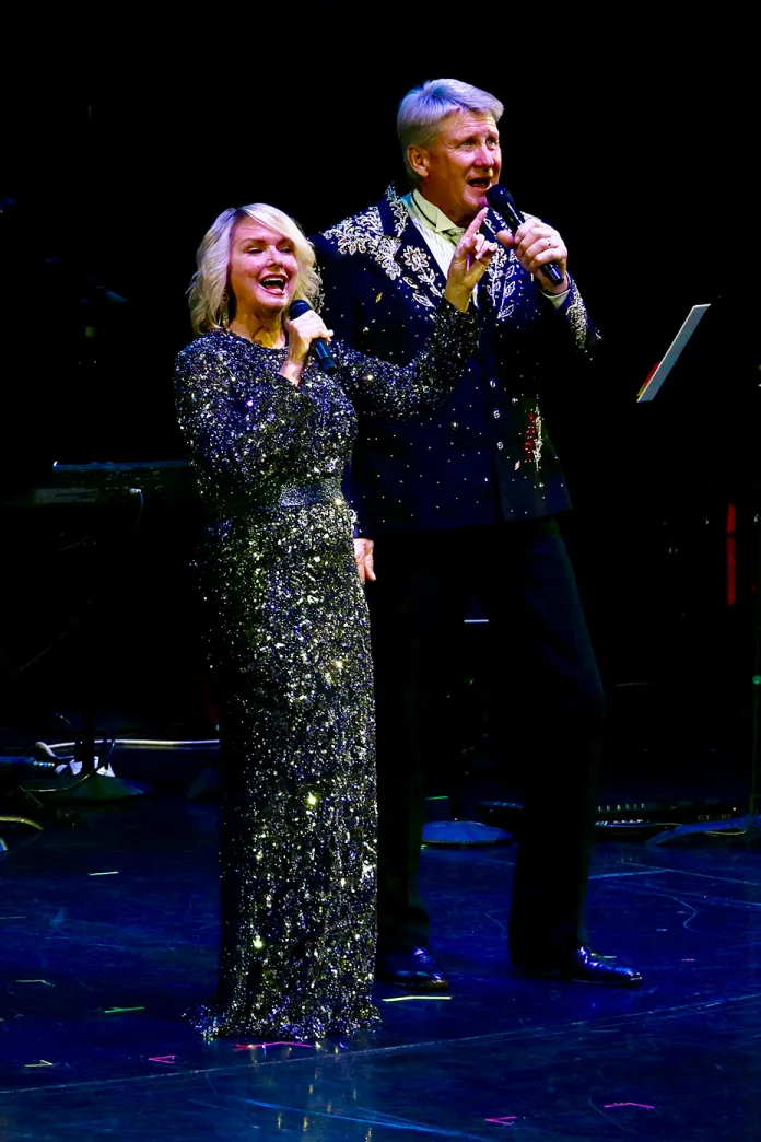 Two singers in glittering outfits perform onstage with microphones under blue lights.