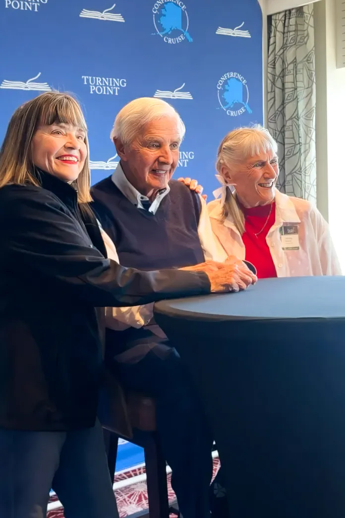 David Jeremiah and two travelers smiling at a tall table before a blue sponsor backdrop.