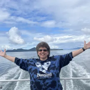 Smiling woman with arms out, standing on a ship deck with the ocean in the background