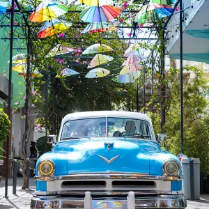 Light blue vintage car under rows of small, colorful umbrellas