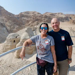 Two travelers smiling at a scenic overlook near the Qumran caves in Israel, with desert cliffs and rocky hills in the background.