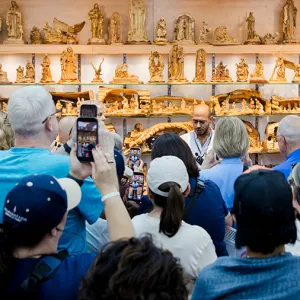 A crowd of people gathers around a vendor booth displaying rows of carved figurines and nativity scenes on wooden shelves, while several attendees hold up phones to take photos.