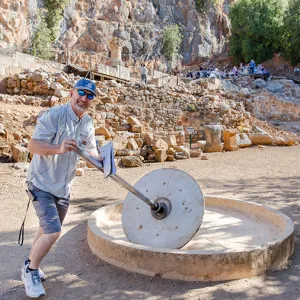 A man in sunglasses and a blue cap demonstrates an ancient stone grinding wheel at an outdoor archaeological site surrounded by rocky ruins and seated visitors in the background.